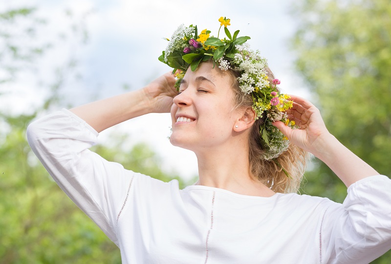 Beautiful young blond woman with flower  wreath on the meadow on a warm summer day. Smiling girl with wild flowers wreath enjoying sun outdoors. Happy woman in the garden. Lifestyle, summer concept